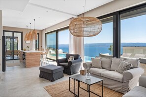 Een moderne woonkamer met een grote bank, een salontafel en een eethoek met een bar. De kamer heeft grote ramen met uitzicht op zee.