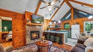 A cozy cabin interior with a stone fireplace, wooden walls, a dining area with bar stools, and a kitchen with a refrigerator.