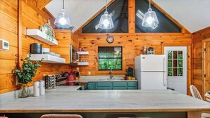 A kitchen with wooden walls, a marble countertop, and a large island with bar stools. There are hanging lights, shelves with plants, and a refrigerator.