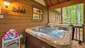 A wooden cabin with a hot tub, a wicker chair with towels, and a window overlooking a forest.
