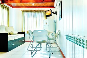 Ein kleines Zimmer mit Bett, einem Tisch mit Stühlen, einem Kühlschrank und einem an der Wand befestigten Fernseher.