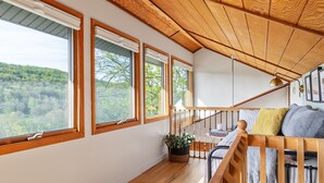 Ein gemütliches Zimmer mit einer Holztreppe, großen Fenstern mit Blick auf einen bewaldeten Hang und einer bequemen Sitzecke mit Kissen.