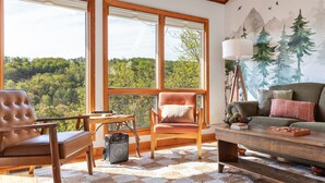 Ein gemütliches Wohnzimmer mit einem großen Fenster, einem Holztisch, einem Ledersessel und einer Couch mit Kissen.
