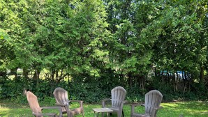 Een paar Adirondack stoelen staan rond een kleine tafel in een grasveld, met een dichte boompartij op de achtergrond.