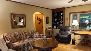 Een woonkamer met een gestreept sofa, een leren fauteuil, een houten salontafel en een eettafel met een lamp.