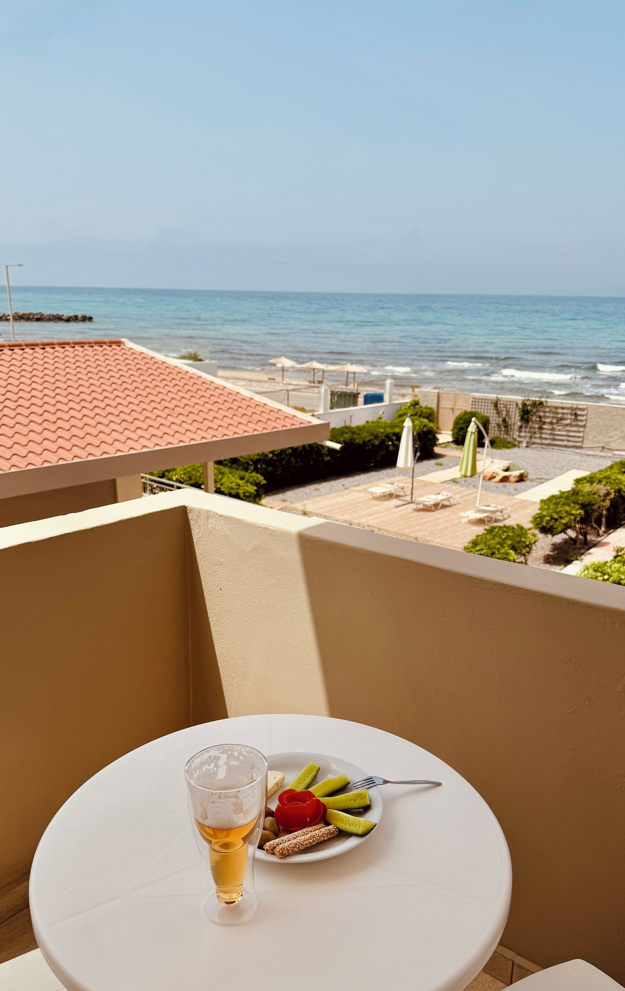 Un balcón con una mesa, una cerveza y un plato de comida. Se ve el océano y una playa con sombrillas.