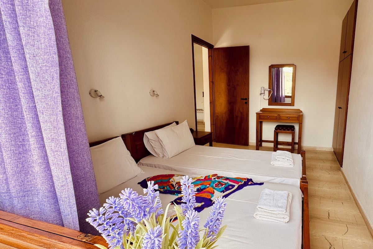 Une chambre d’hôtel avec deux lits, une commode en bois, un miroir et une porte donnant sur une autre pièce.