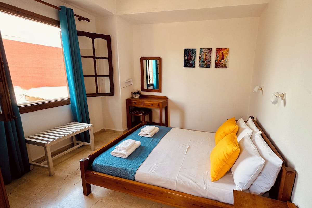 Une chambre d’hôtel avec un lit en bois, un banc, un bureau et une fenêtre avec des rideaux.
