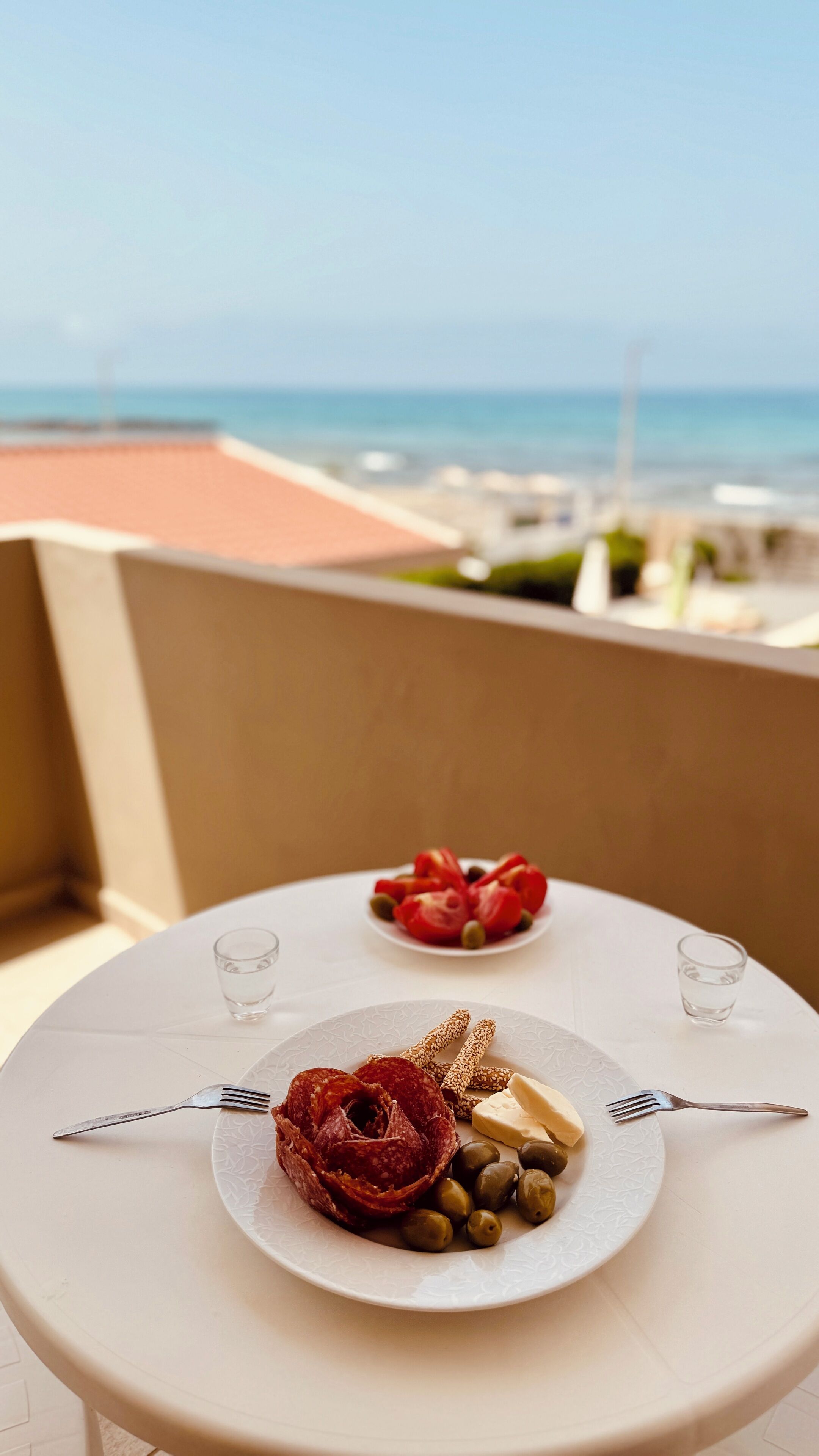 Two tables with plates of food and glasses of water, set on a balcony with a view of the sea.