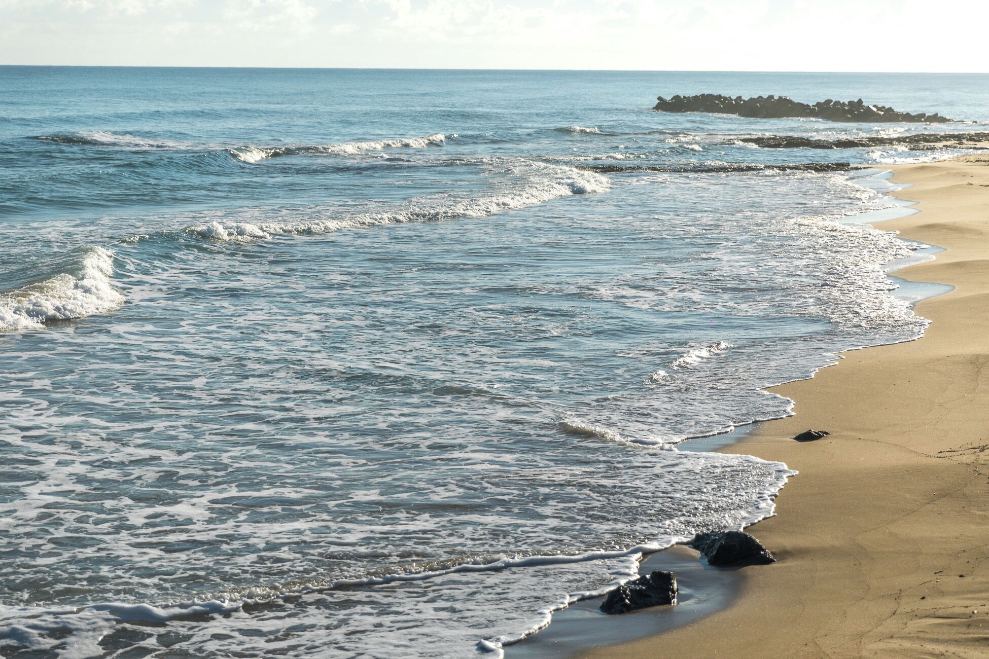 A beach with waves gently lapping onto the shore, a clear sky, and a distant rocky outcrop.