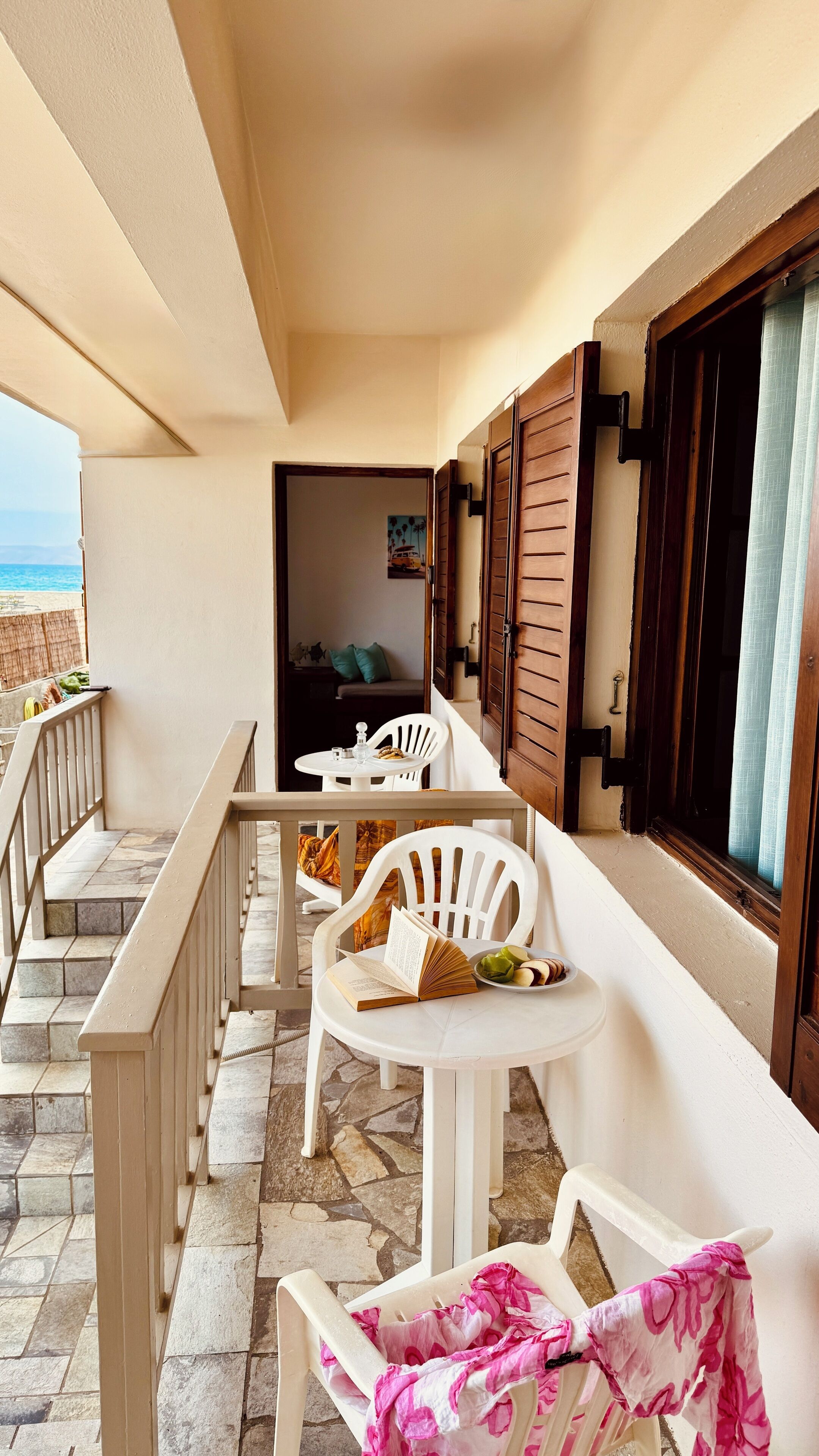 Un balcón con sillas blancas, una mesa con un libro y aperitivos, y vista al mar.