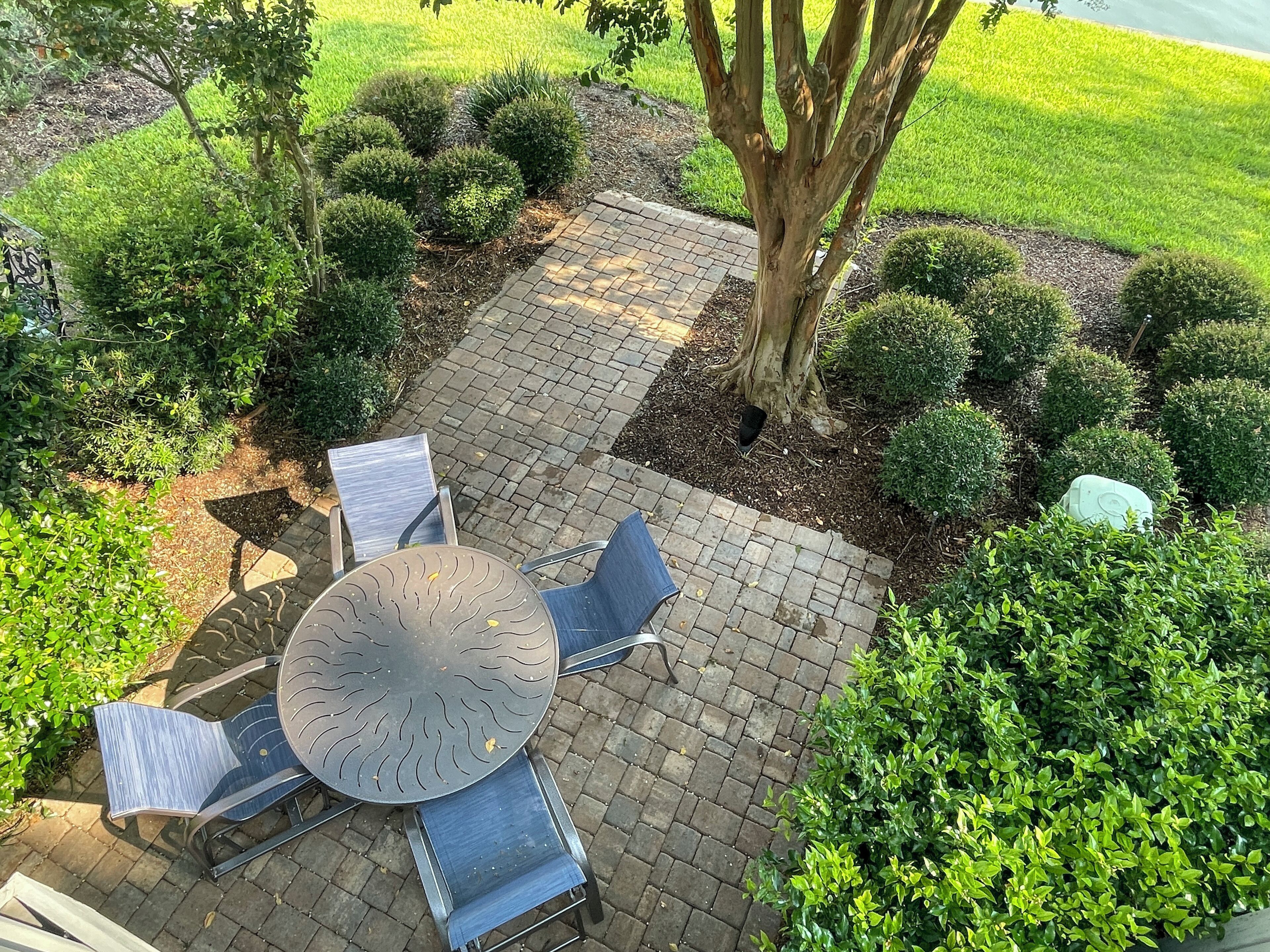 A patio area with a round table and four chairs, surrounded by a brick pathway and lush greenery.