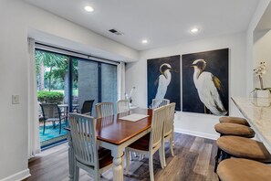 A dining area with a wooden table and white chairs, a large painting of birds on the wall, and a view through the glass door to an outdoor patio. - 490 Plantation Club Villas at The Sea Pines Resort (Hilton Head Island)