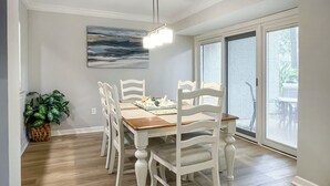 Un comedor con mesa de madera y sillas blancas, una planta en maceta y un cuadro en la pared.