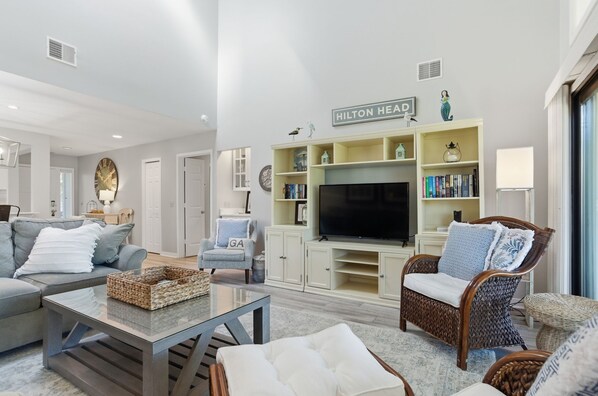 A modern living room with a flat-screen TV, a sofa, a wicker chair, and a bookshelf. - 563 Ocean Course at The Sea Pines Resort (Hilton Head Island)