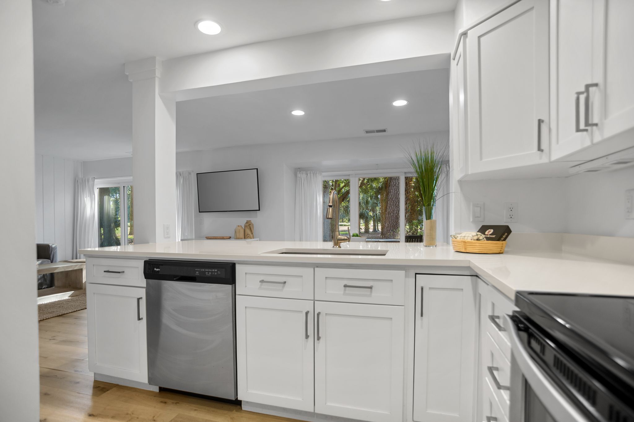 Una cocina moderna con armarios blancos, lavavajillas, estufa y pileta. Se observa una ventana con cortinas y una planta en la encimera.