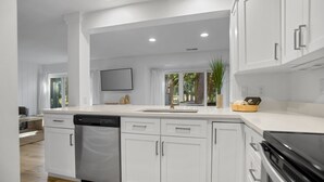 A modern kitchen with white cabinetry, a dishwasher, a stove, and a sink. There is a window with curtains and a plant on the counter.