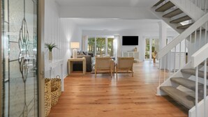A spacious living room with a wooden floor, white walls, a staircase, and a flat-screen TV mounted on the wall.