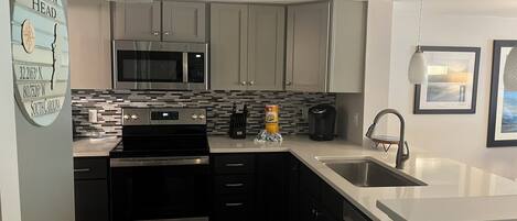 A modern kitchen with stainless steel appliances, a microwave, and a coffee maker. There is a sink, a stove, and a dishwasher. The kitchen has a tiled backsplash and light-colored cabinets.
