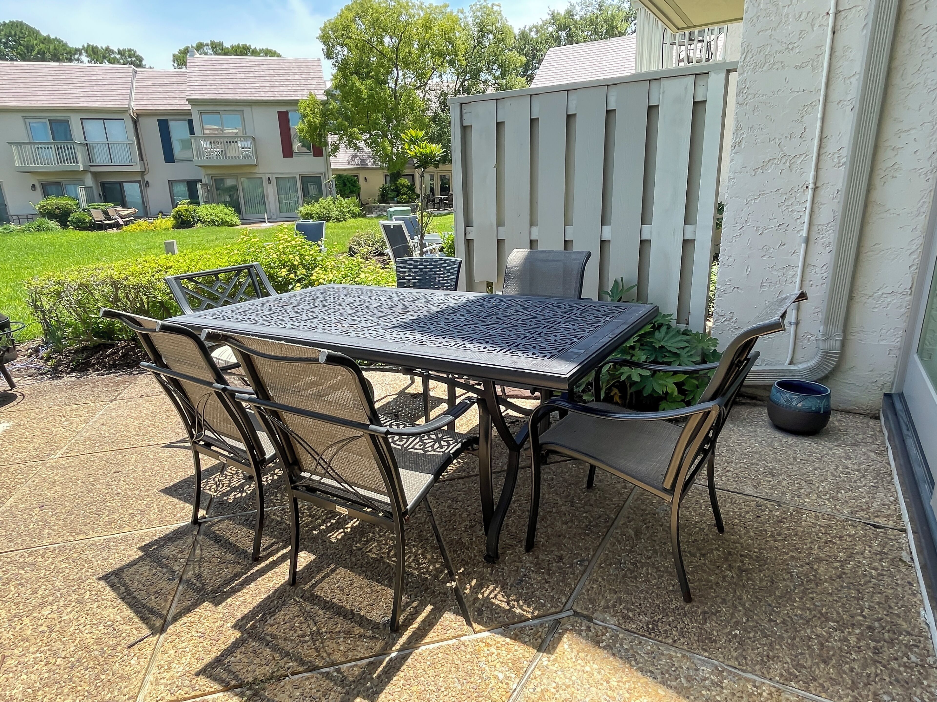 Un patio al aire libre con una mesa y sillas de metal, rodeado por una valla blanca y follaje.