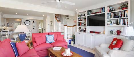Amplia sala de estar con un sofá rojo, un sofá blanco y una mesa de centro de madera. Hay una estantería llena de libros y objetos decorativos, un televisor de pantalla plana montado en la pared y un ventilador de techo.