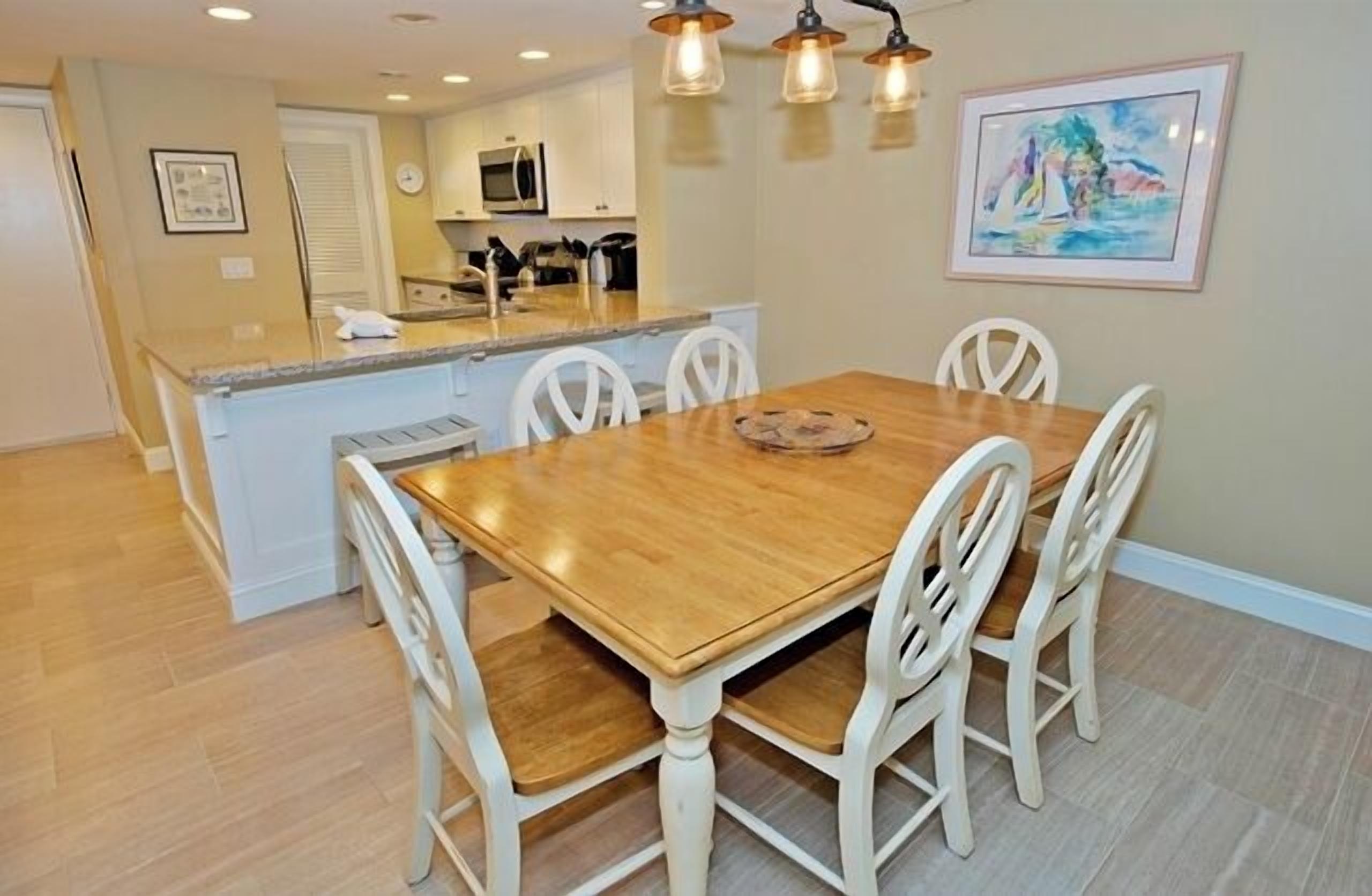 A dining area with a wooden table and white chairs, a kitchen in the background, and a painting on the wall.