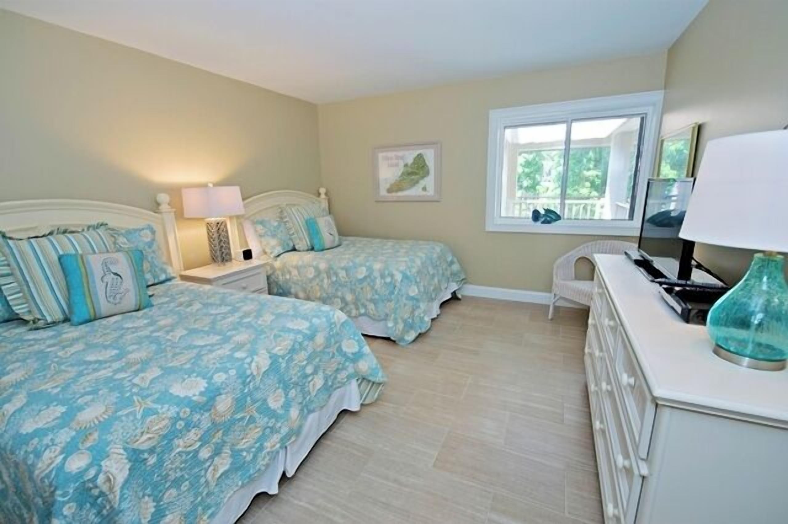 A bedroom with two beds, a nightstand with a lamp, a dresser, and a window with a view of greenery.