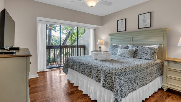 Une chambre à coucher comprenant un lit, un ventilateur de plafond, une table de chevet, une commode et une fenêtre donnant sur des arbres.