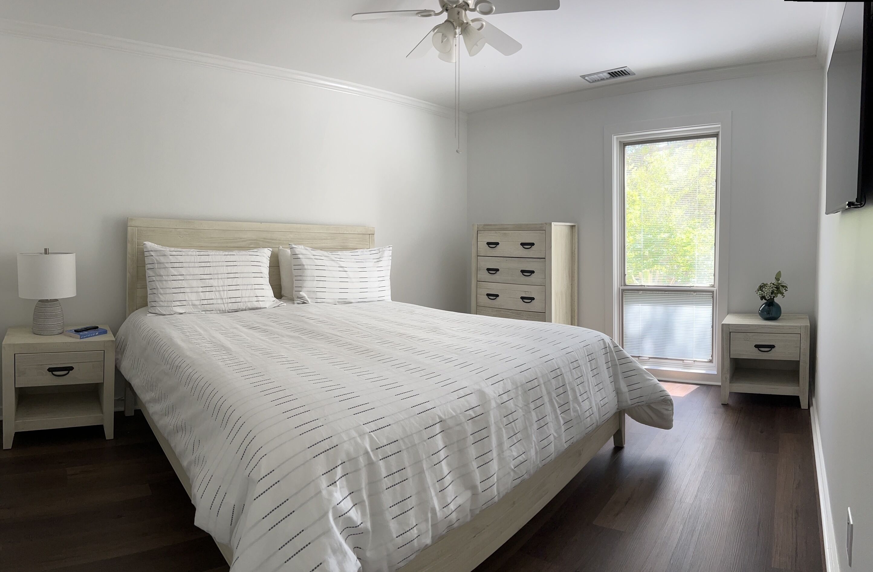 Une chambre à coucher avec un lit, des tables de chevet, un ventilateur de plafond, une fenêtre et une petite plante.