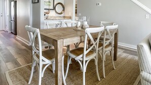 A dining area with a wooden table and white chairs, a kitchen in the background, and a staircase leading upstairs.