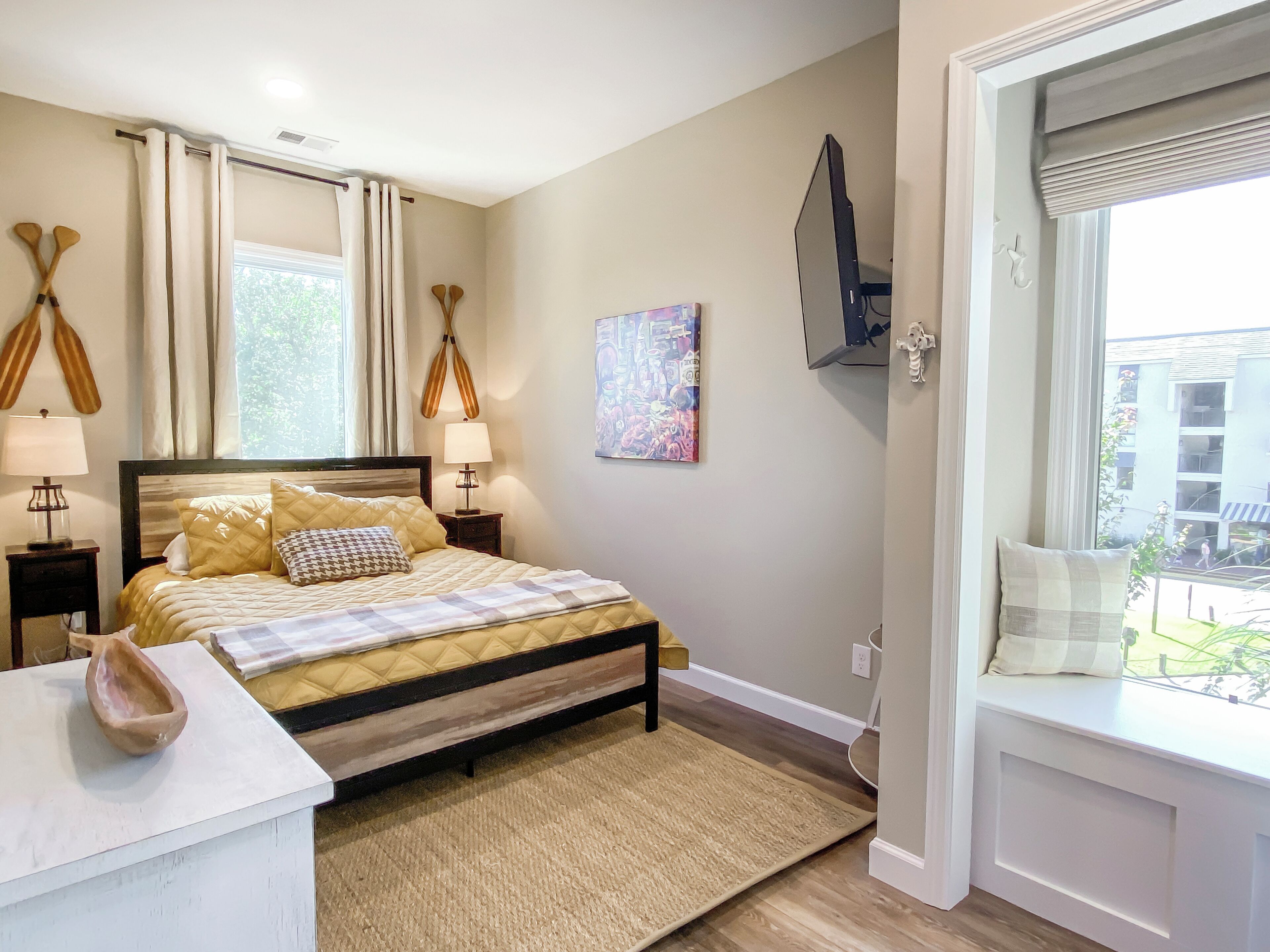 A neatly arranged bedroom with a bed, bedside lamps, a painting, and a view of the outside.