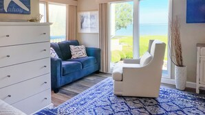 A room with a white dresser, a blue sofa, a white armchair, and a large window with a view of the sea.