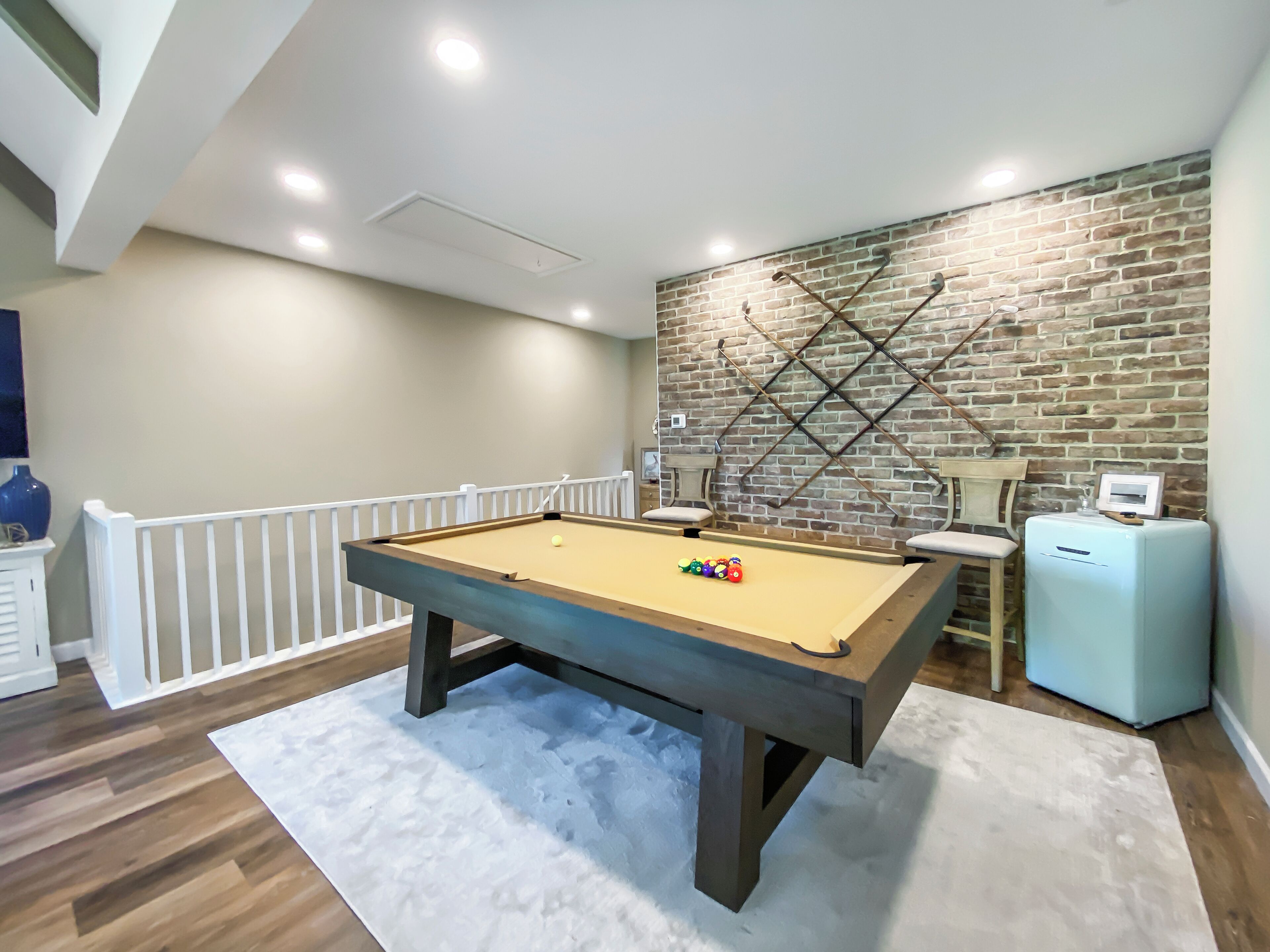 A pool table in a room with a brick wall, white railing, and a small table with a lamp.