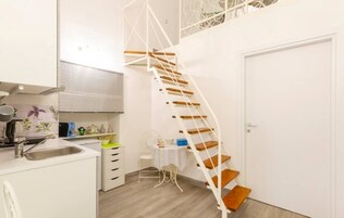 Una cocina moderna con gabinetes blancos, un lavamanos y un comedor con mesa y sillas. Hay una puerta blanca y una escalera con escalones de madera.