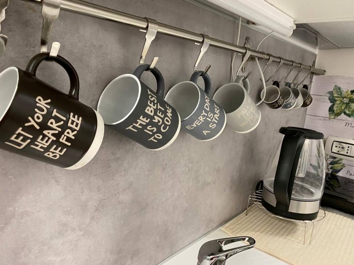 A kitchen with a coffee machine, a kettle, and a series of mugs hanging on a rack with motivational quotes.