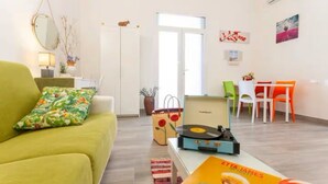 A living room with a green sofa, a coffee table with a record player, a dining area with a table and chairs, and a door leading to another room. - Iris Apartment - comfort e stile (Bisceglie)