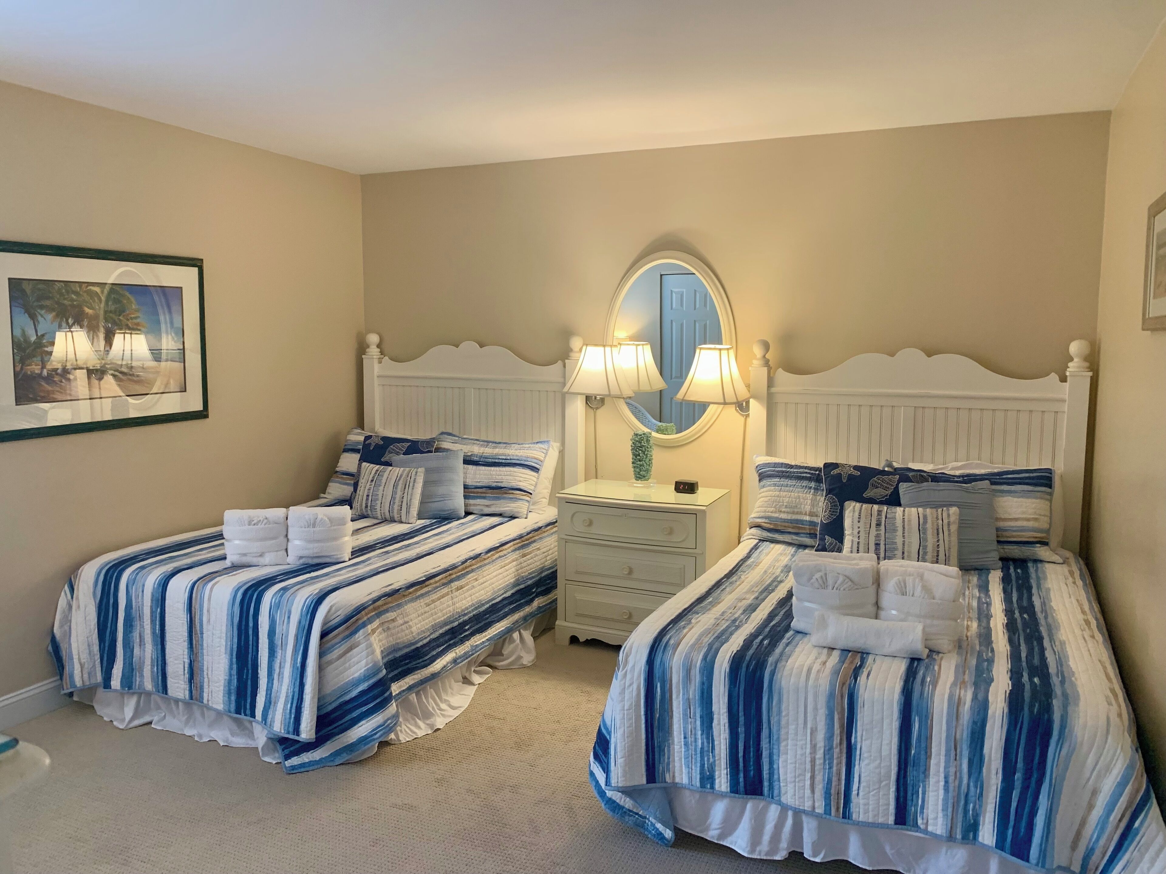 Une chambre d’hôtel avec deux lits, chacun doté de draps et d’oreillers à rayures, une table de chevet avec une lampe, un miroir et un tableau encadré accroché au mur.