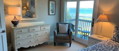 A bedroom with a bed, a dresser, a chair, a nightstand, a lamp, a mirror, and a view of the ocean through a sliding door.