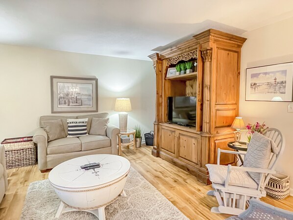 Un salon avec un canapé beige, une table basse blanche, un meuble en bois sur lequel se trouve un téléviseur, et un tableau encadré accroché au mur.