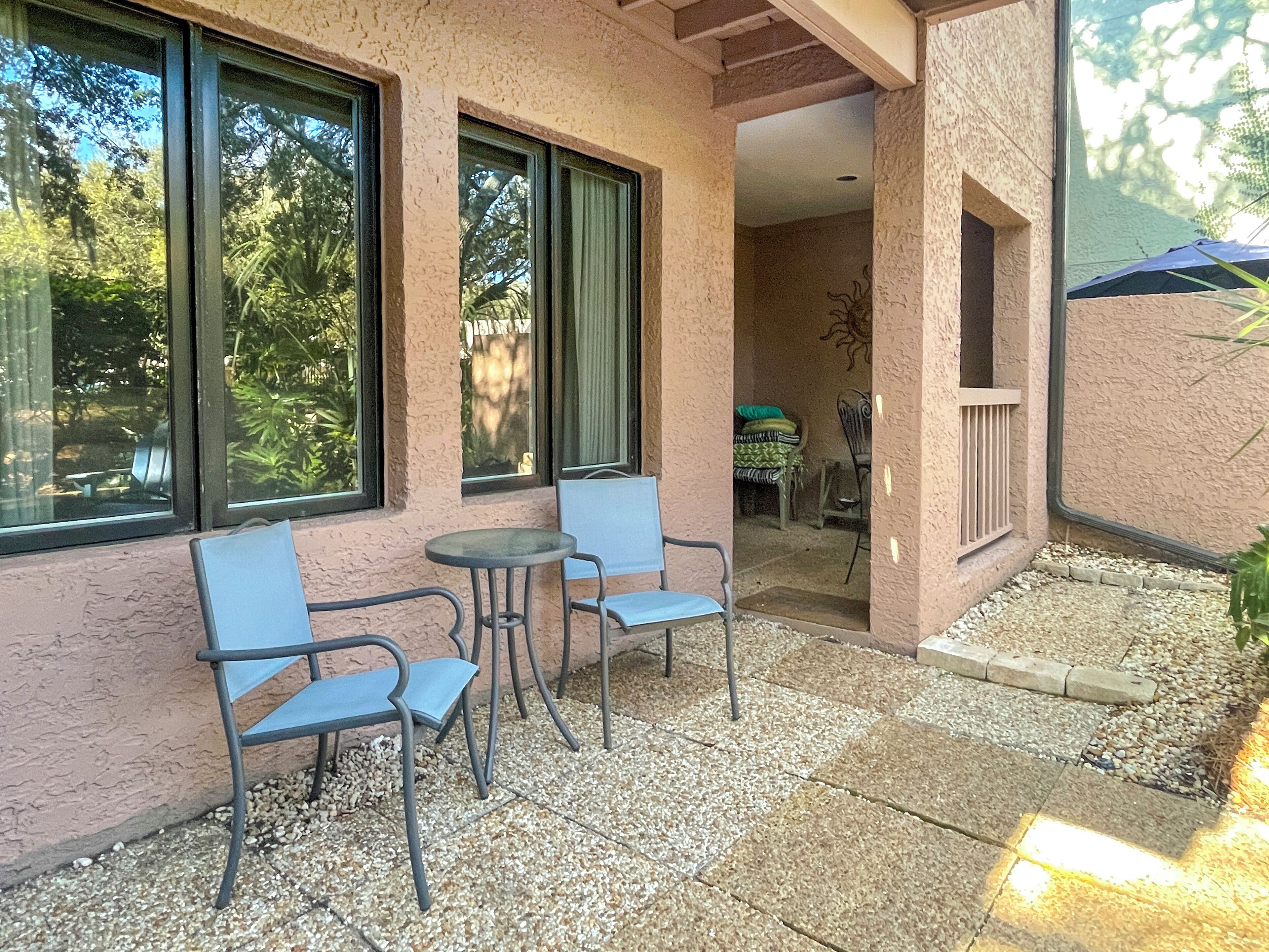 Eine Terrasse mit zwei Stühlen und einem Tisch, ein überdachter Bereich mit Grill und Blick auf Bäume durch große Fenster.