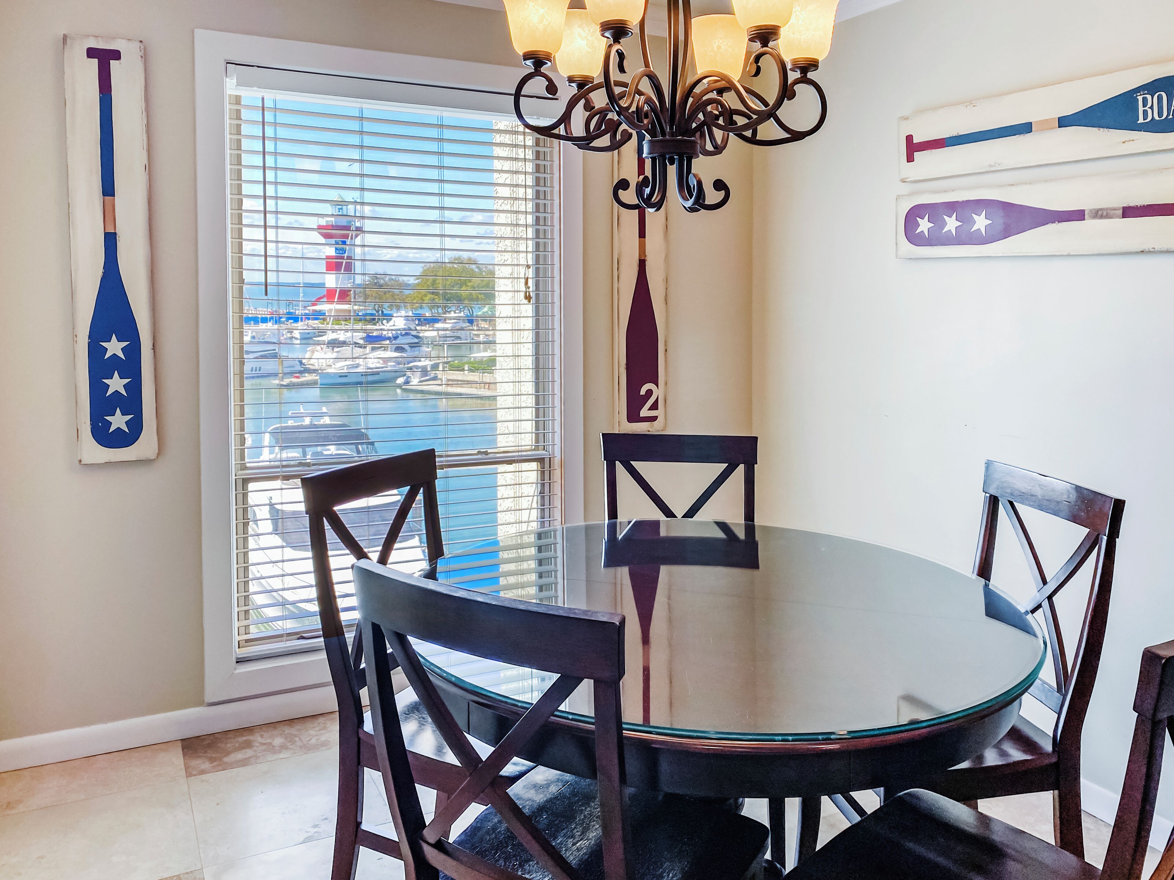 A dining area with a round glass table, four wooden chairs, a chandelier, and a view of a marina through a window with blinds.