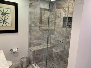 A bathroom with a glass-enclosed shower, marble tiles, a framed artwork, and a toilet with a trash can. - 841 Ketch Court at The Sea Pines Resort (Hilton Head Island)