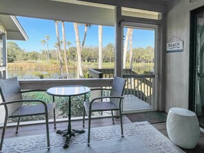 A covered outdoor seating area with two chairs and a table, overlooking a lake and palm trees. - 564 Ocean Course at The Sea Pines Resort (Hilton Head Island)
