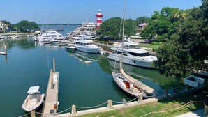 A marina with multiple yachts docked, a lighthouse in the background, and lush greenery surrounding the area.