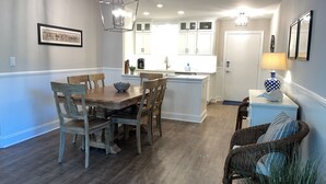 A dining area with a wooden table and chairs, a kitchen with white cabinets, and a wicker chair with a cushion. - 880 Ketch Court at The Sea Pines Resort (Hilton Head Island)