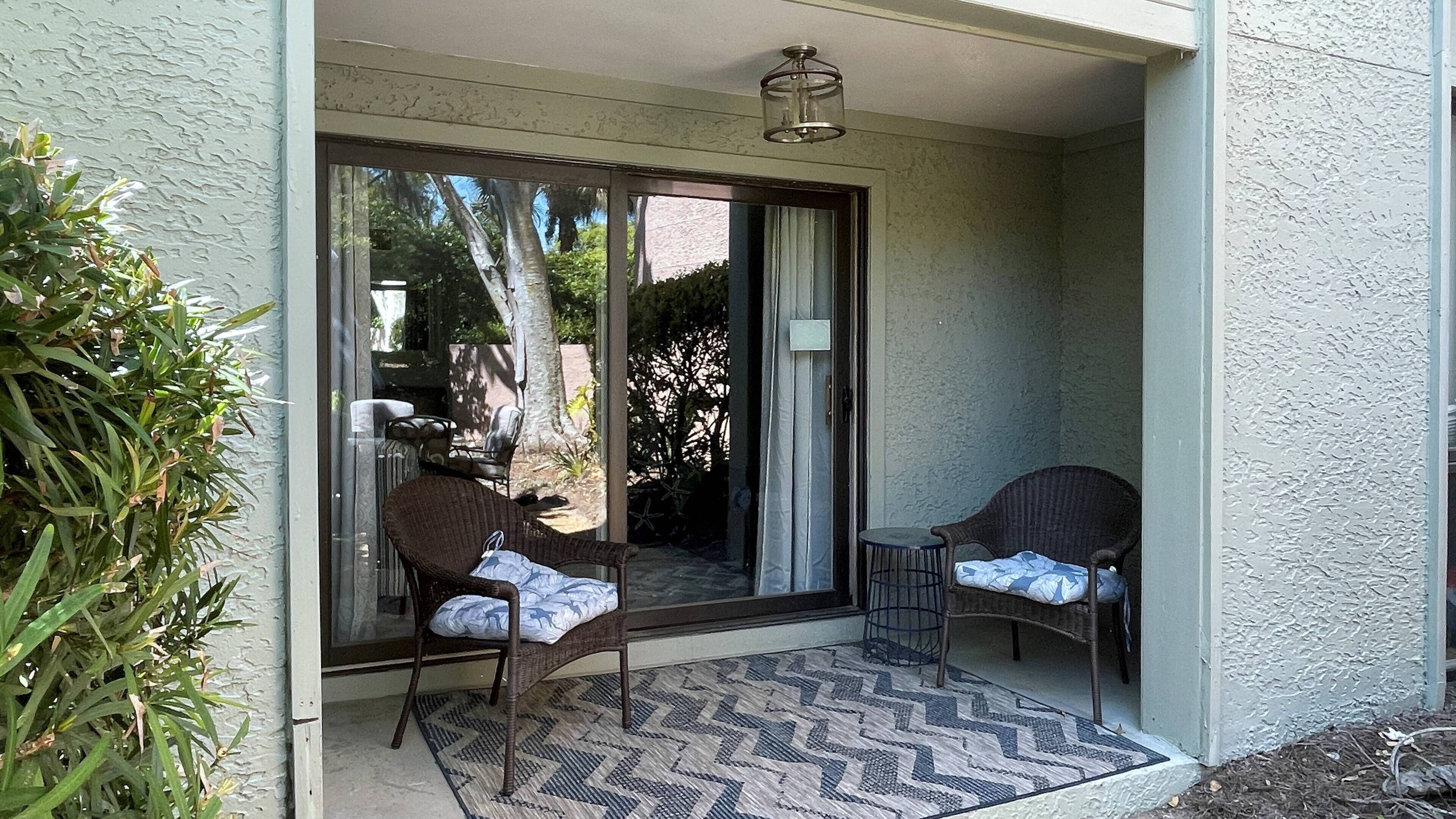 A covered patio with two wicker chairs and a small table, a sliding glass door leading to a garden, and a textured wall.