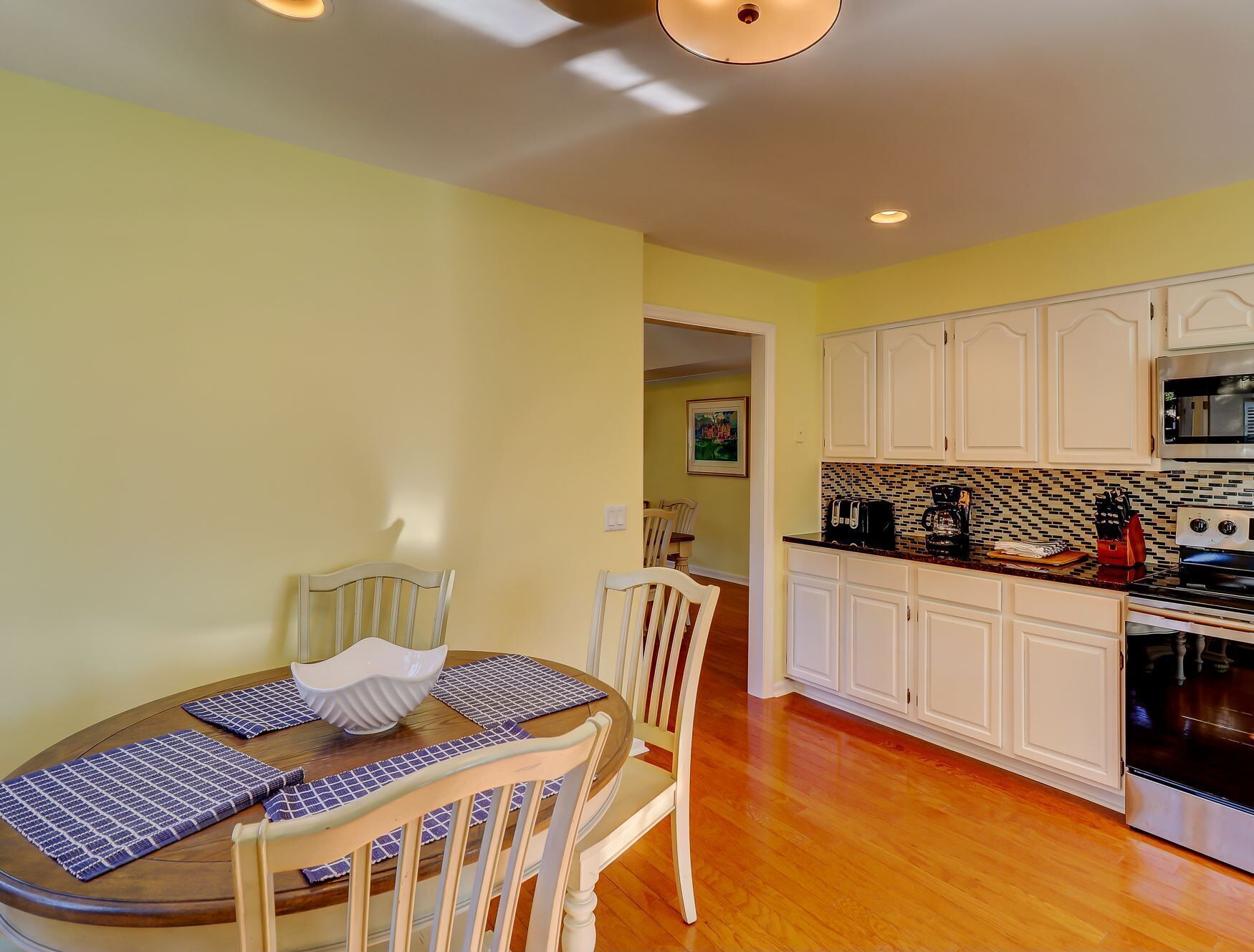 A dining area with a round table, chairs, and a kitchen with white cabinets and a black microwave.