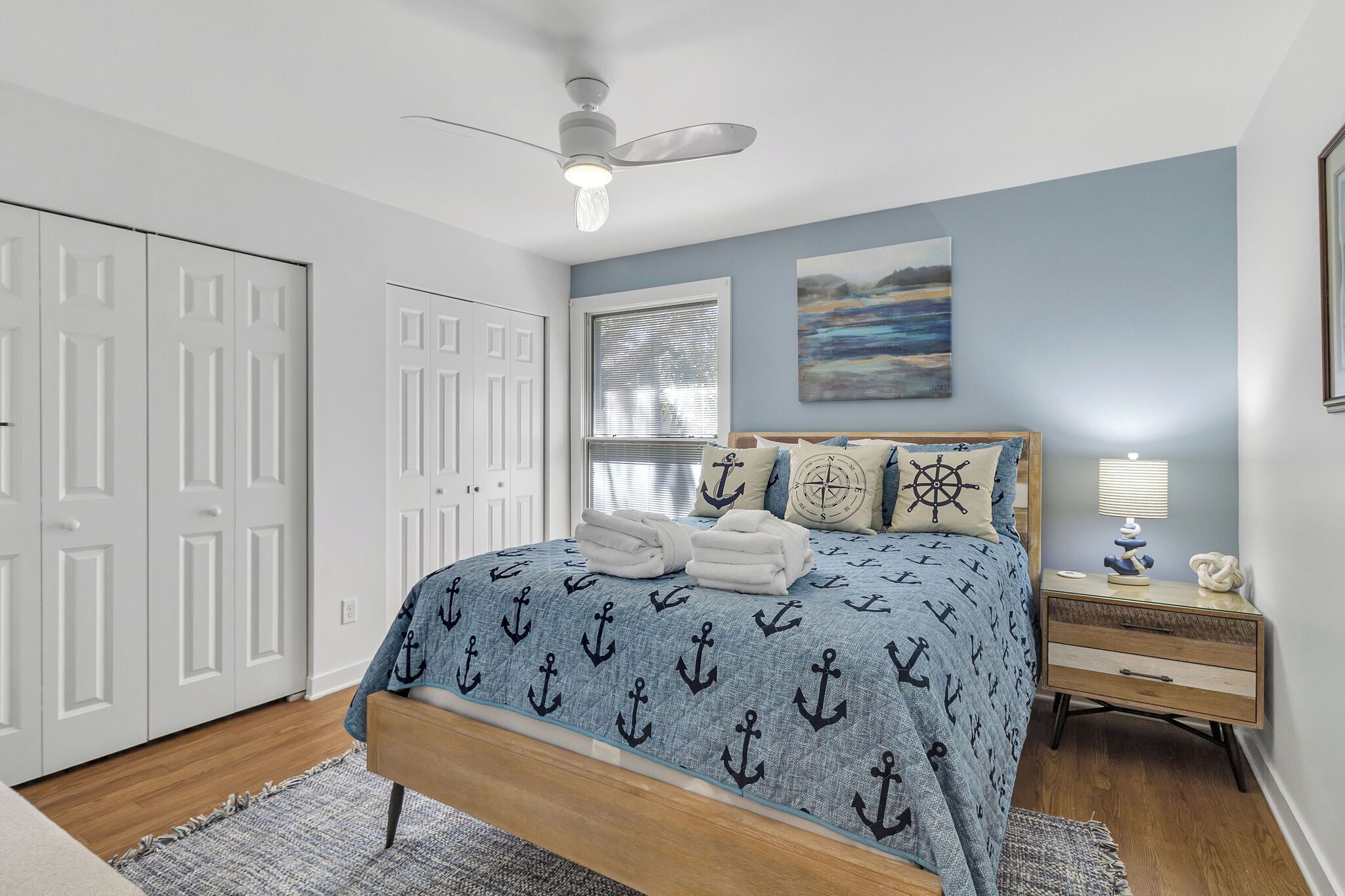 A bedroom with a bed, a nightstand, a ceiling fan, a window with blinds, and a door.