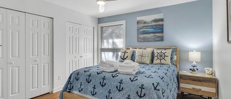 A bedroom with a bed, a nightstand, a ceiling fan, a window with blinds, and a door.