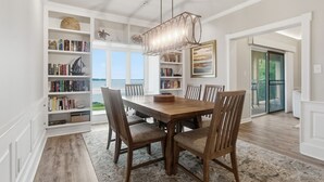 Un comedor con una mesa y sillas de madera, rodeado de estanterías, una araña y vistas al exterior.